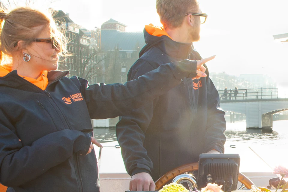 A group of people enjoying drinks on a boat with a view of a river and a bridge in the background during sunset.