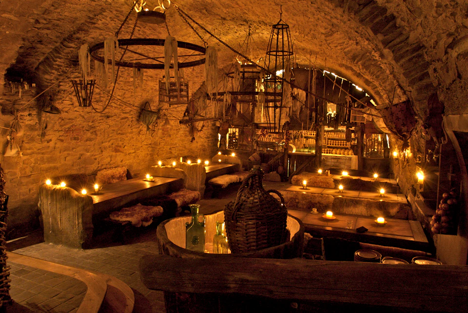 Dimly lit rustic tavern with stone walls, wooden tables, sheepskin-covered benches, candles, and a bottle-filled bar in the background.