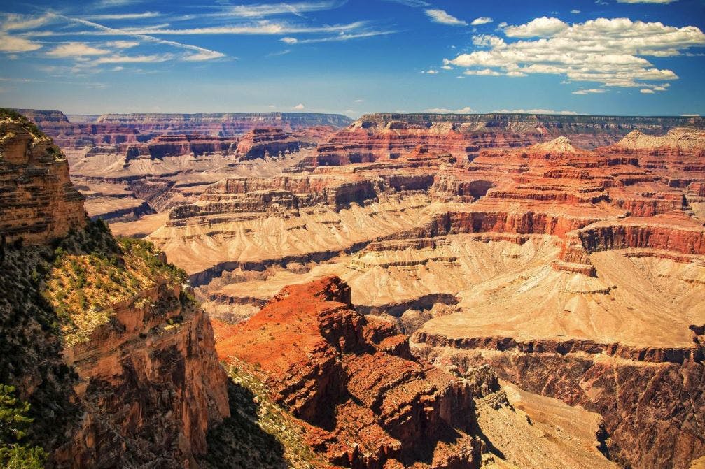 Expansive view of the Grand Canyon with layered red and brown rock formations under a partly cloudy blue sky.