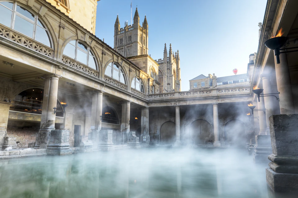 A historic bathhouse with steamy water, surrounded by columns and arches, with a cathedral visible in the background.