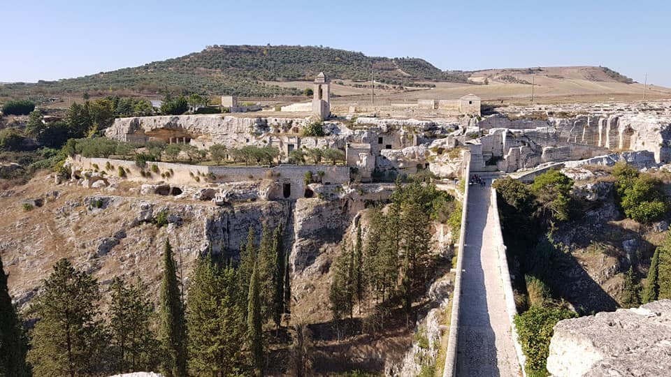 Old stone bridge leading to a historic village with stone buildings, trees, and hills in the background under a clear blue sky.