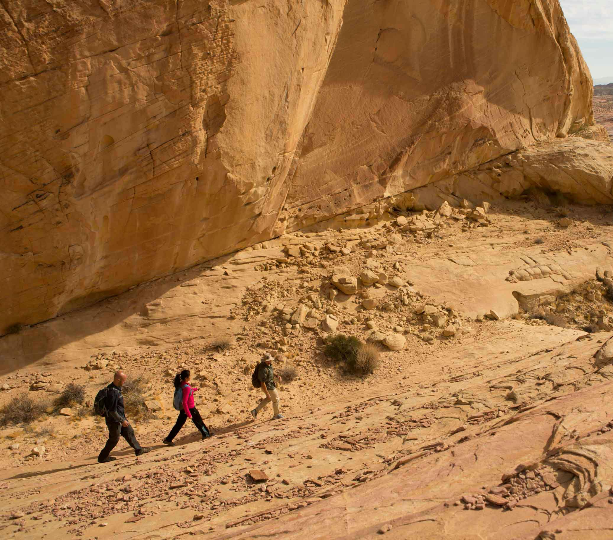 Three people hike along the rocky slope of a large desert cliff under a clear sky.