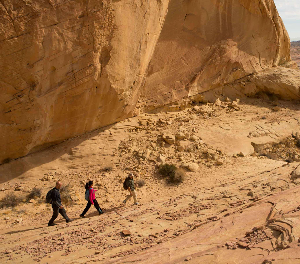 Three people hike along the rocky slope of a large desert cliff under a clear sky.