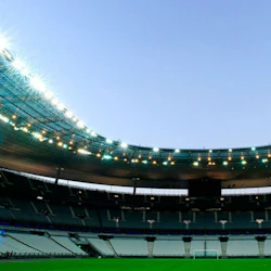 An empty, large stadium with tiered seating and bright lights, showing a green field and clear sky above.