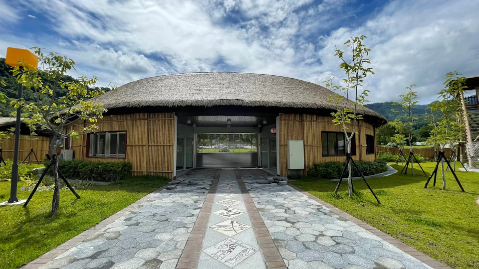 A building with a thatched roof and bamboo walls, featuring a patterned walkway leading to its entrance, under a cloudy sky.