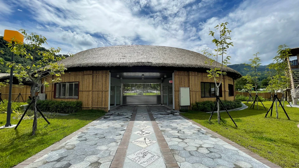 A building with a thatched roof and bamboo walls, featuring a patterned walkway leading to its entrance, under a cloudy sky.