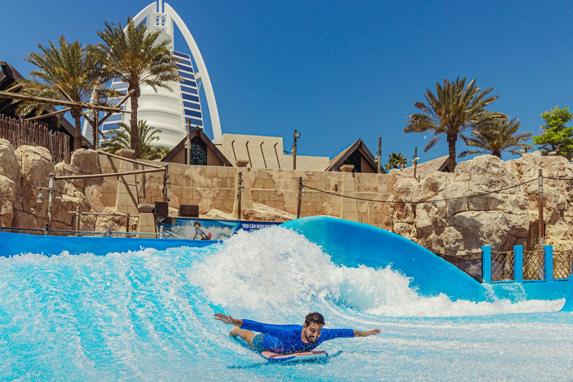 Un homme portant un rash guard bleu surfe sur une vague en bodyboard dans un parc aquatique, avec des palmiers et un bâtiment en arrière-plan.