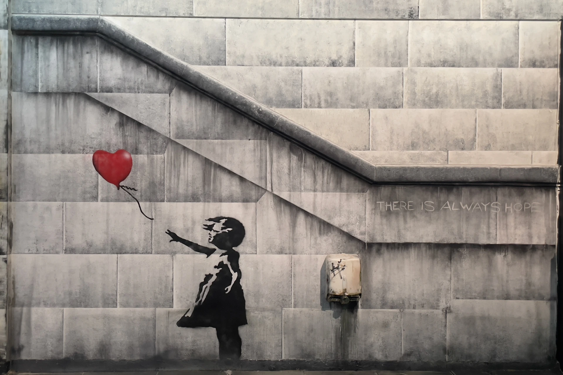 A graffiti of a girl reaching for a red heart-shaped balloon is painted on a grey wall with a staircase and the words "THERE IS ALWAYS".