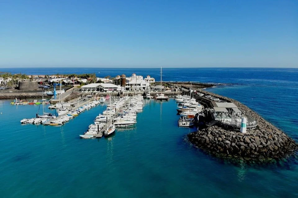 Marina with numerous boats docked in clear blue water, surrounded by buildings and rocky breakwaters under a clear blue sky.
