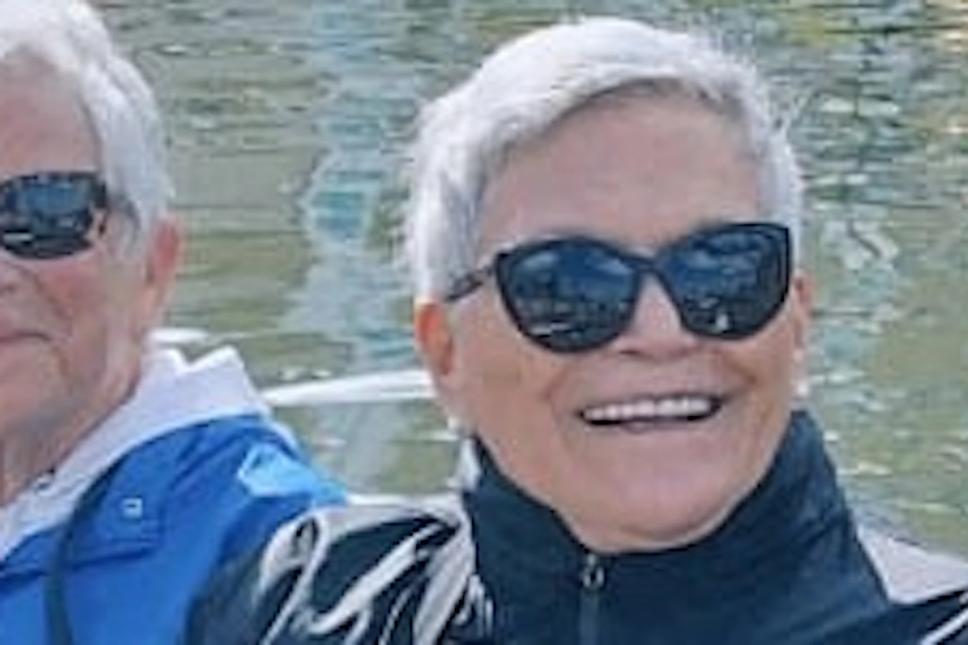 Four women wearing sunglasses and jackets sit on a boat docked by a canal, with colorful buildings and boats in the background.
