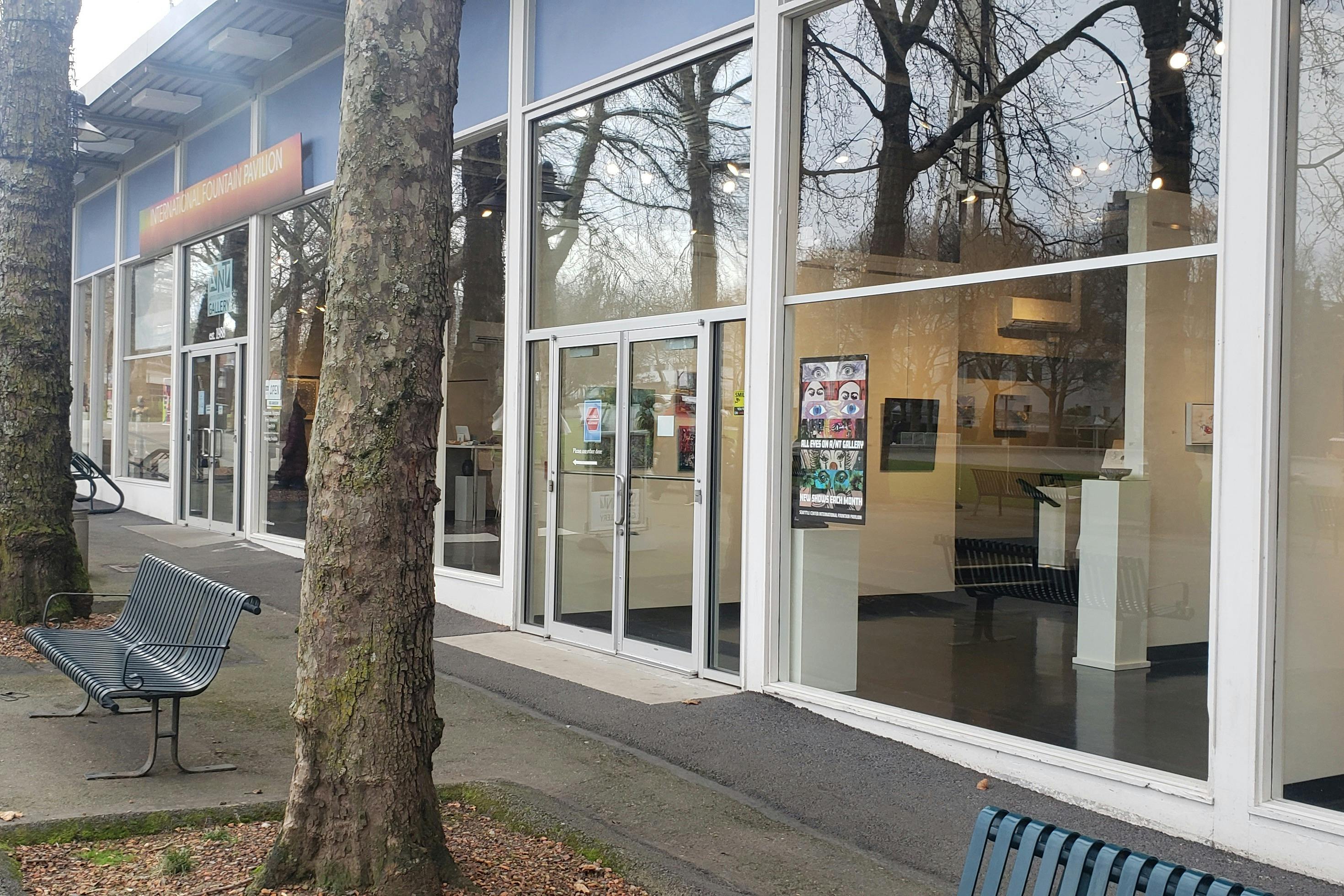 大きな窓にアートワーク、目に見える木々、ベンチが飾られた店舗。 ドアにはポスターがあり、アートギャラリーにつながっています。