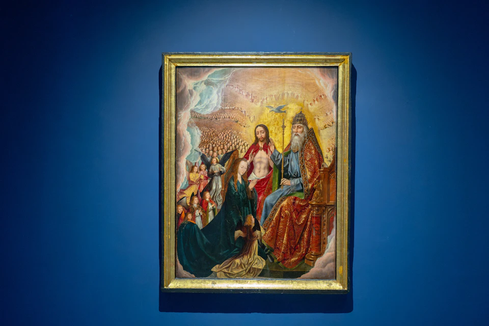 A framed religious artwork displayed on a blue wall, depicting a group of figures and a heavenly scene with vivid colors.
