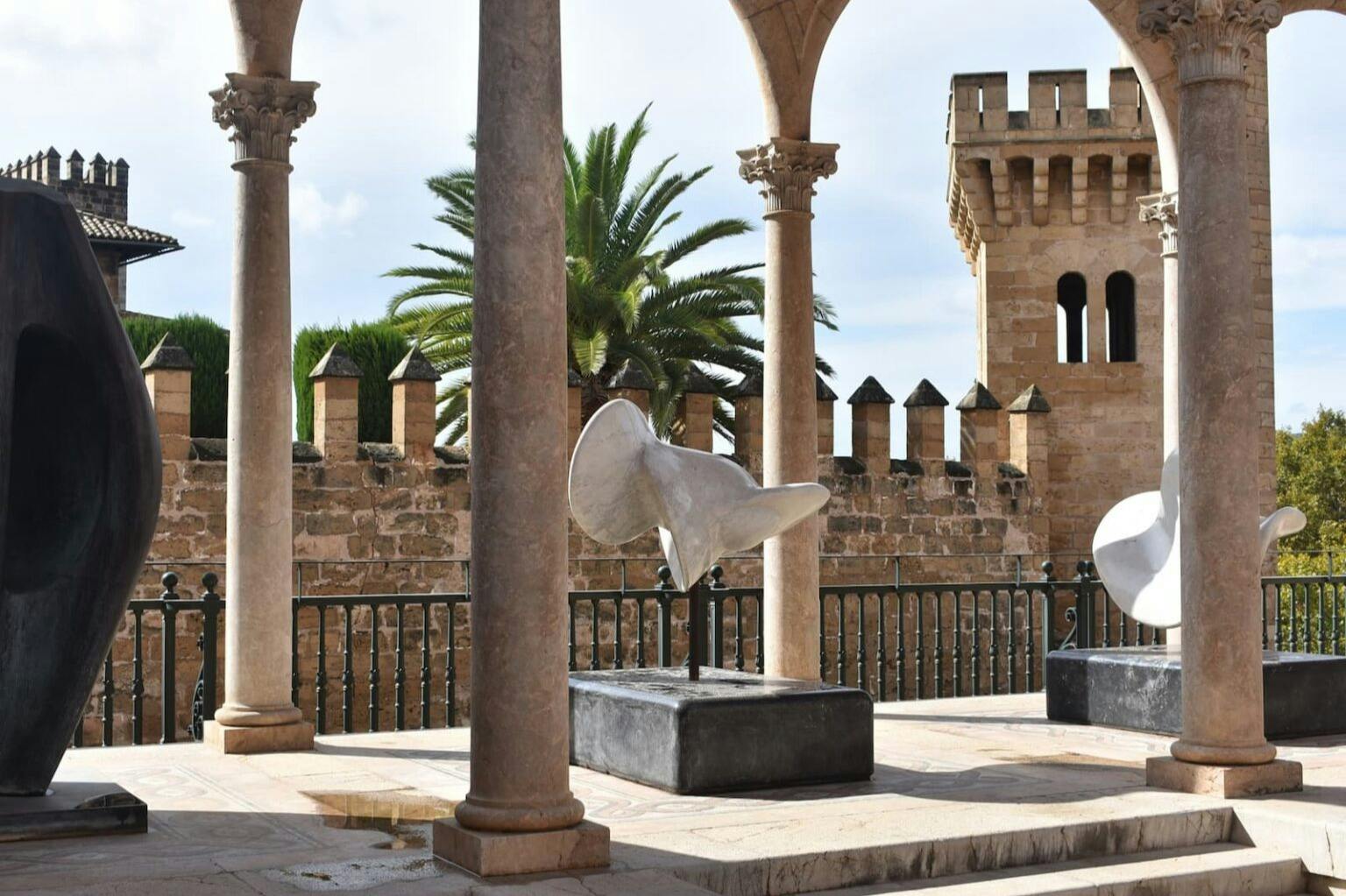 Um pátio com colunas de pedra e esculturas abstractas, encostado a uma muralha de castelo e a palmeiras sob um céu soalheiro.