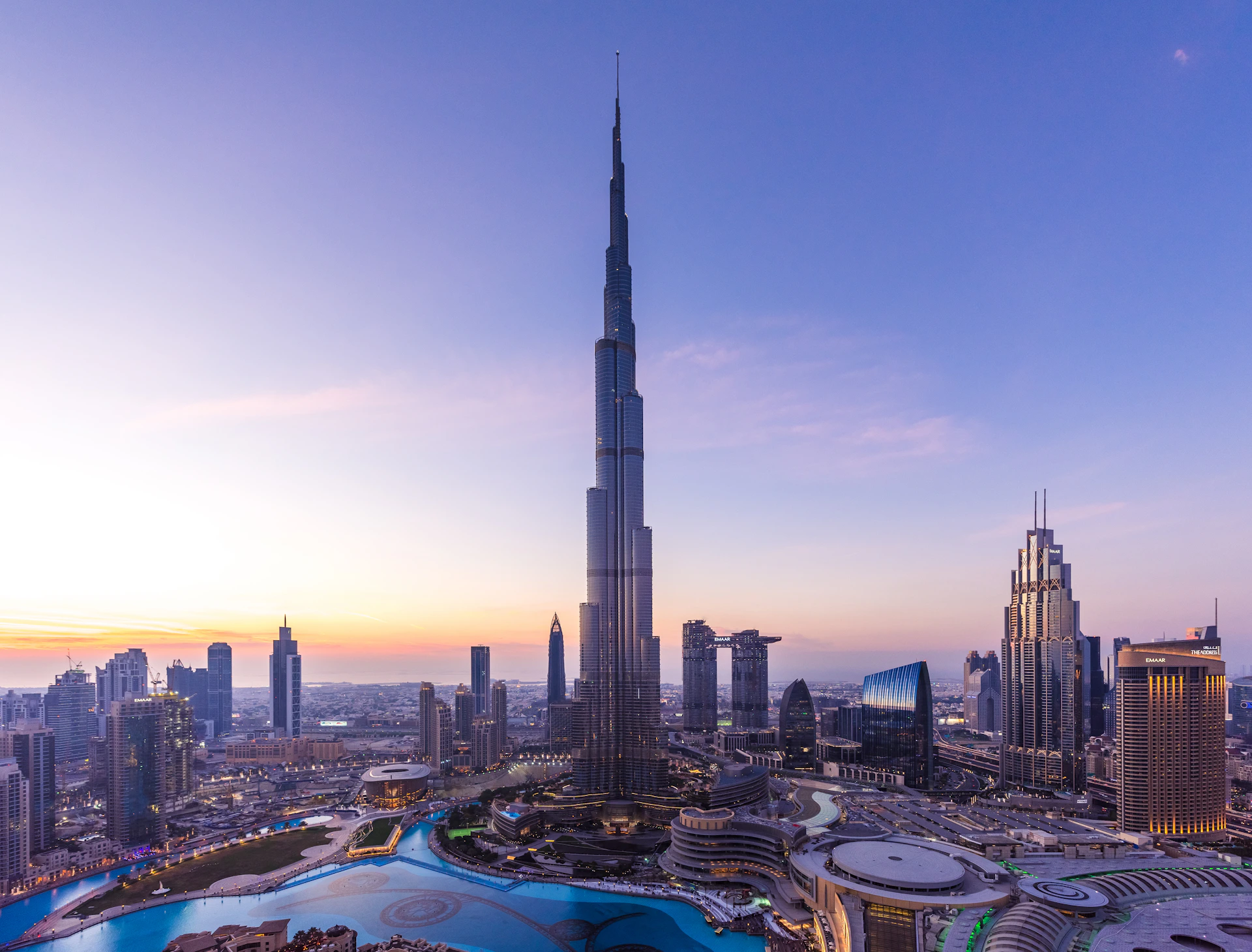 The Burj Khalifa towering over the Dubai skyline at sunset, surrounded by modern buildings and a water fountain.