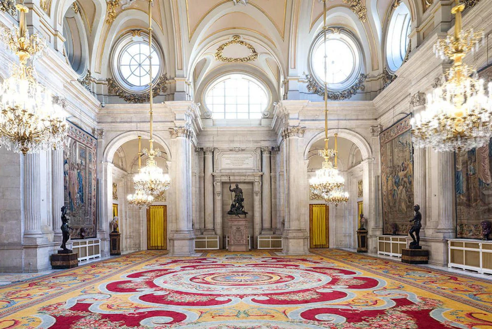 Grand hall with ornate chandeliers, arched windows, a statue, decorative tapestries, and a vibrant patterned carpet.