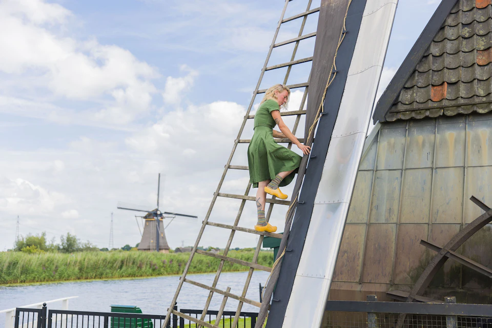 A person in a green dress and yellow clogs climbs a ladder on the side of a windmill, with another windmill in the background.