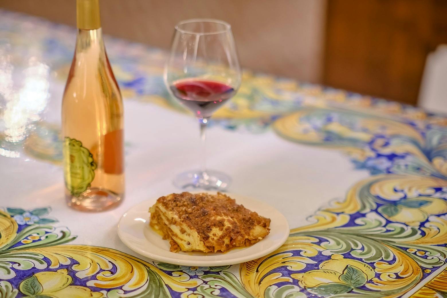 A plate with a portion of lasagna on a table with a colorful tablecloth, next to a glass of red wine and a bottle.