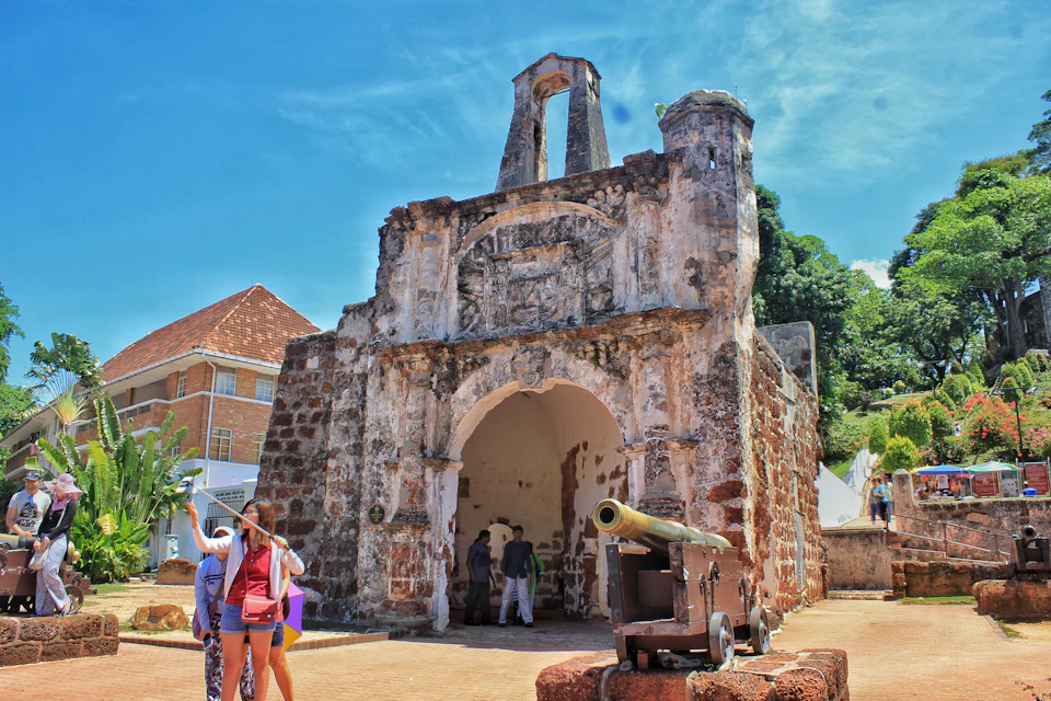 People standing in front of an old, weathered stone archway with a cannon nearby, against a backdrop of trees and a blue sky.