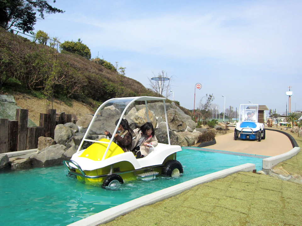 Two people riding in a small, canopy-covered electric vehicle on a narrow water channel in an outdoor park setting.