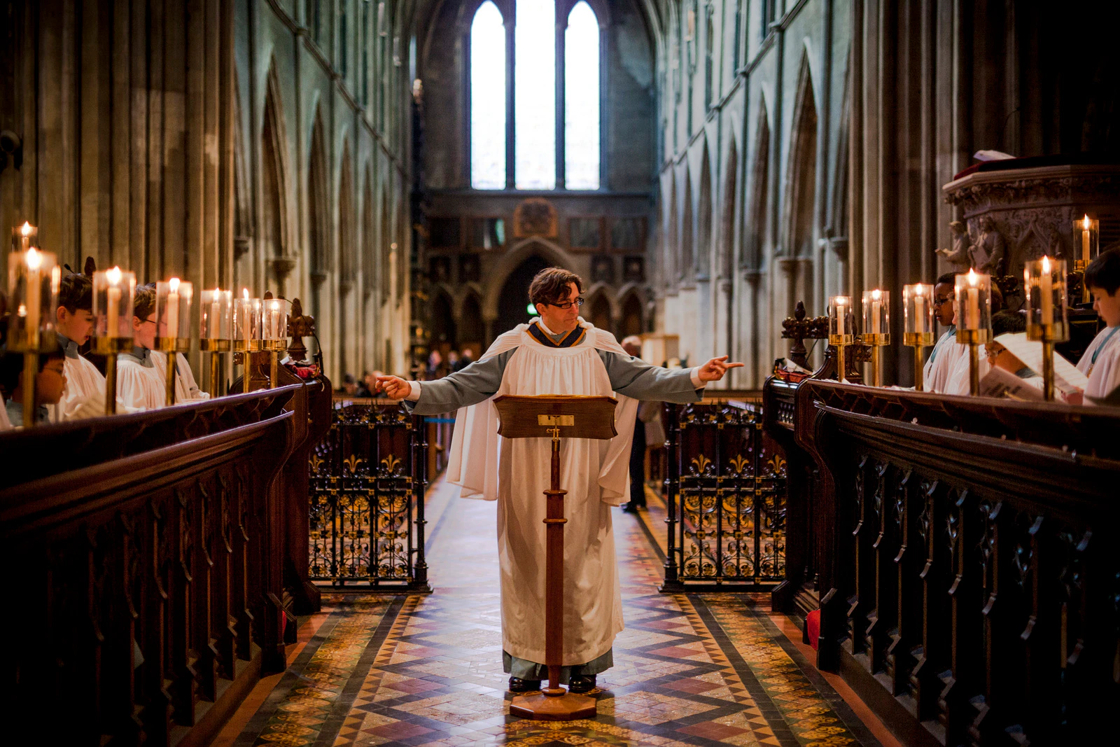 A choir conductor raises his arms in a cathedral, surrounded by candles and ornate decor, with a large arched window behind him.
