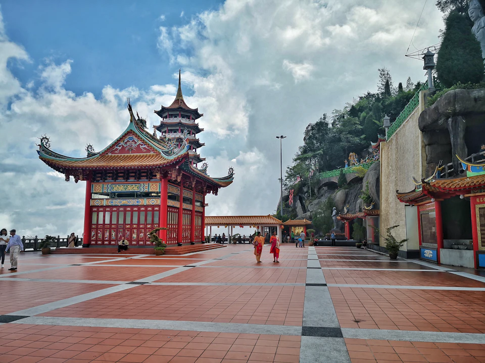 An open courtyard with a red pavilion, a tall pagoda, and people walking. Hillside greenery and cloudy sky in the background.