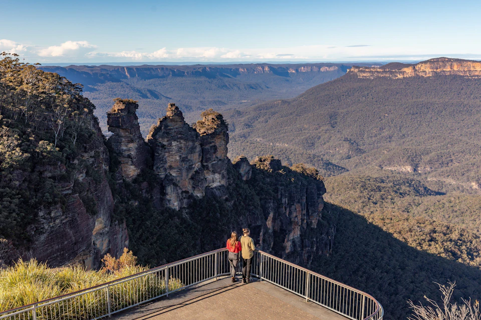 Two people stand on a viewing platform overlooking the Three Sisters rock formation and a vast forested valley.