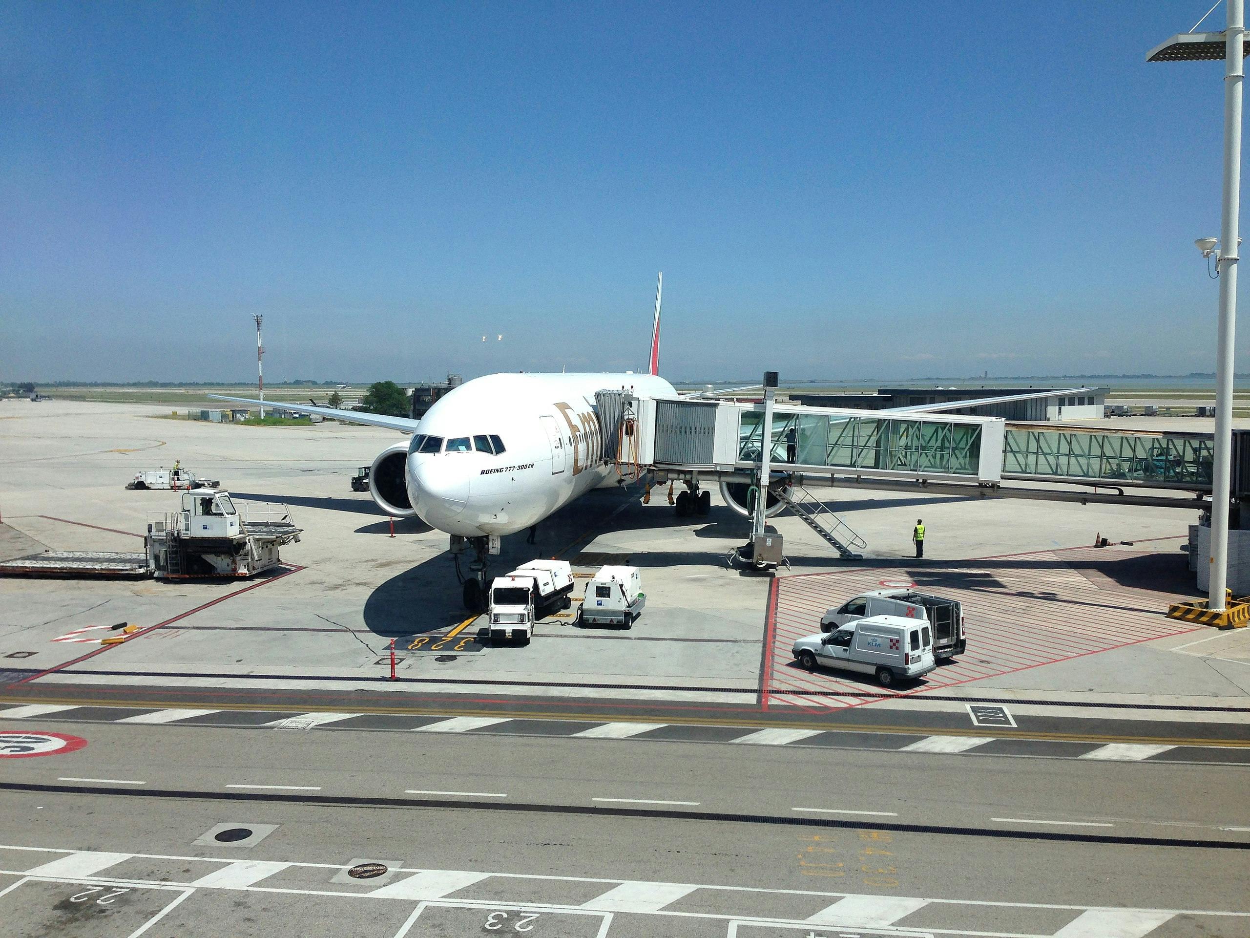 Ένα αεροπλάνο είναι αγκυροβολημένο σε μια πύλη αεροδρομίου με προσαρτημένες γέφυρες τζετ. Οχήματα εξυπηρέτησης και προσωπικό είναι παρόντα στην άσφαλτο.