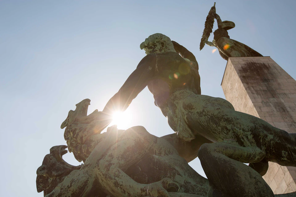 Bronze statues of mythical figures in combat, shot from below with the sun haloing one figure's head, under a clear sky.