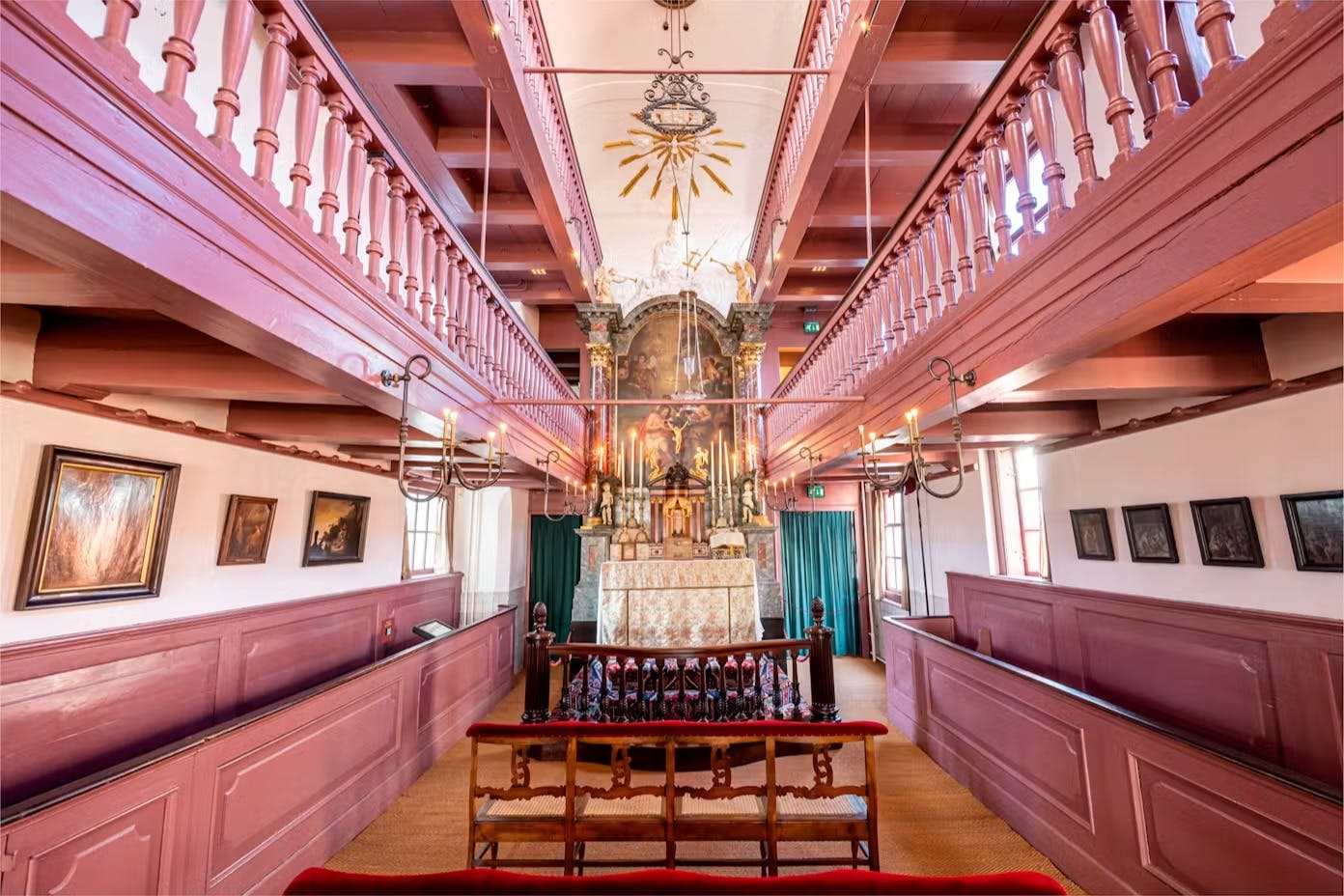 O interior ornamentado de uma capela, com varandas de madeira cor-de-rosa, um altar central com velas e um elemento decorativo no teto.