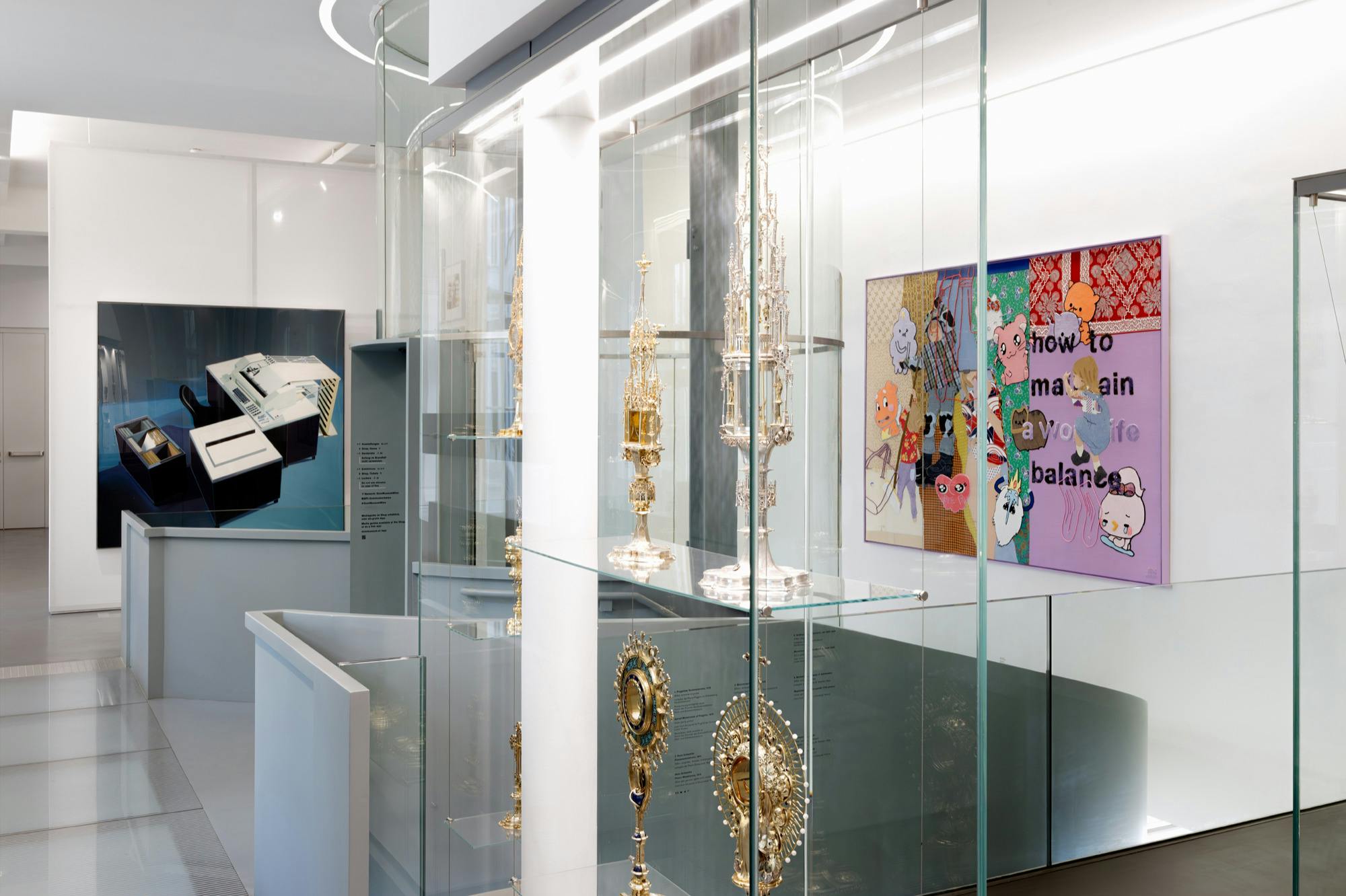 Skleněné vitríny obsahující zdobené kovové artefakty v moderním muzejním prostředí s uměleckými díly na stěnách.
