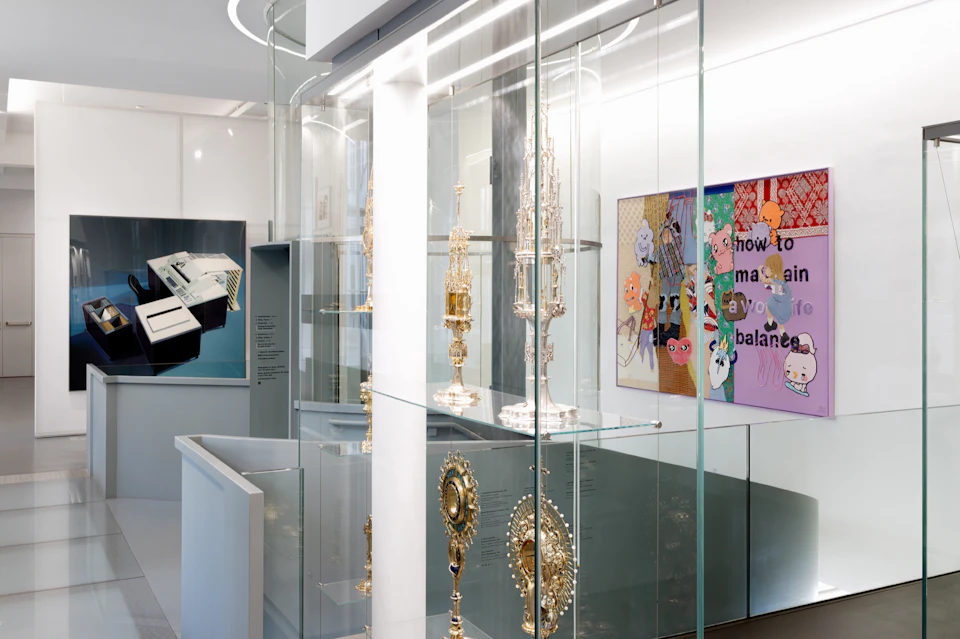 Glass display cases containing ornate metallic artifacts in a modern museum setting, with artwork on the walls.