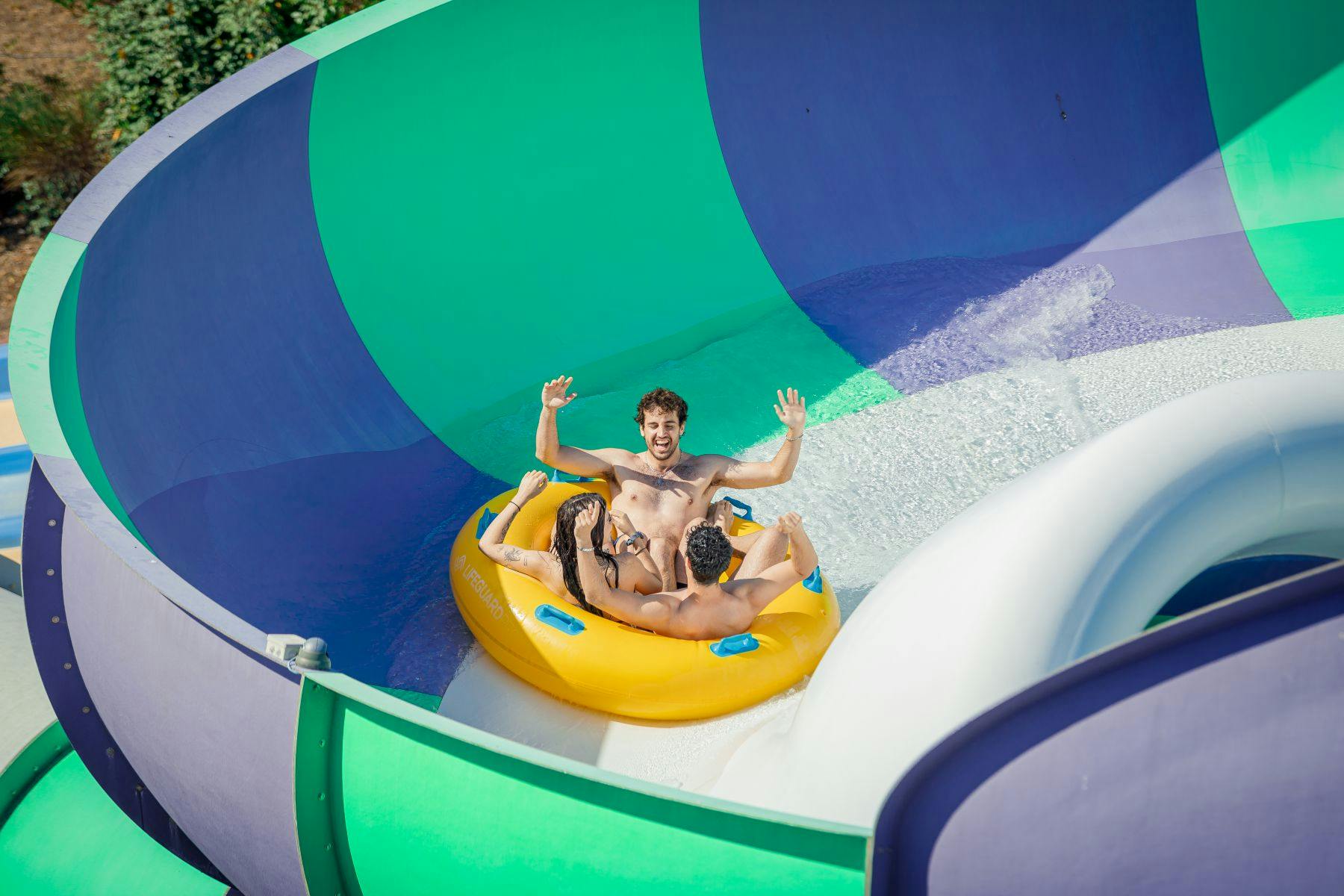 Tres persones en una bassa inflable groga gaudint d'un tobogan d'aigua amb parets corbes de colors.