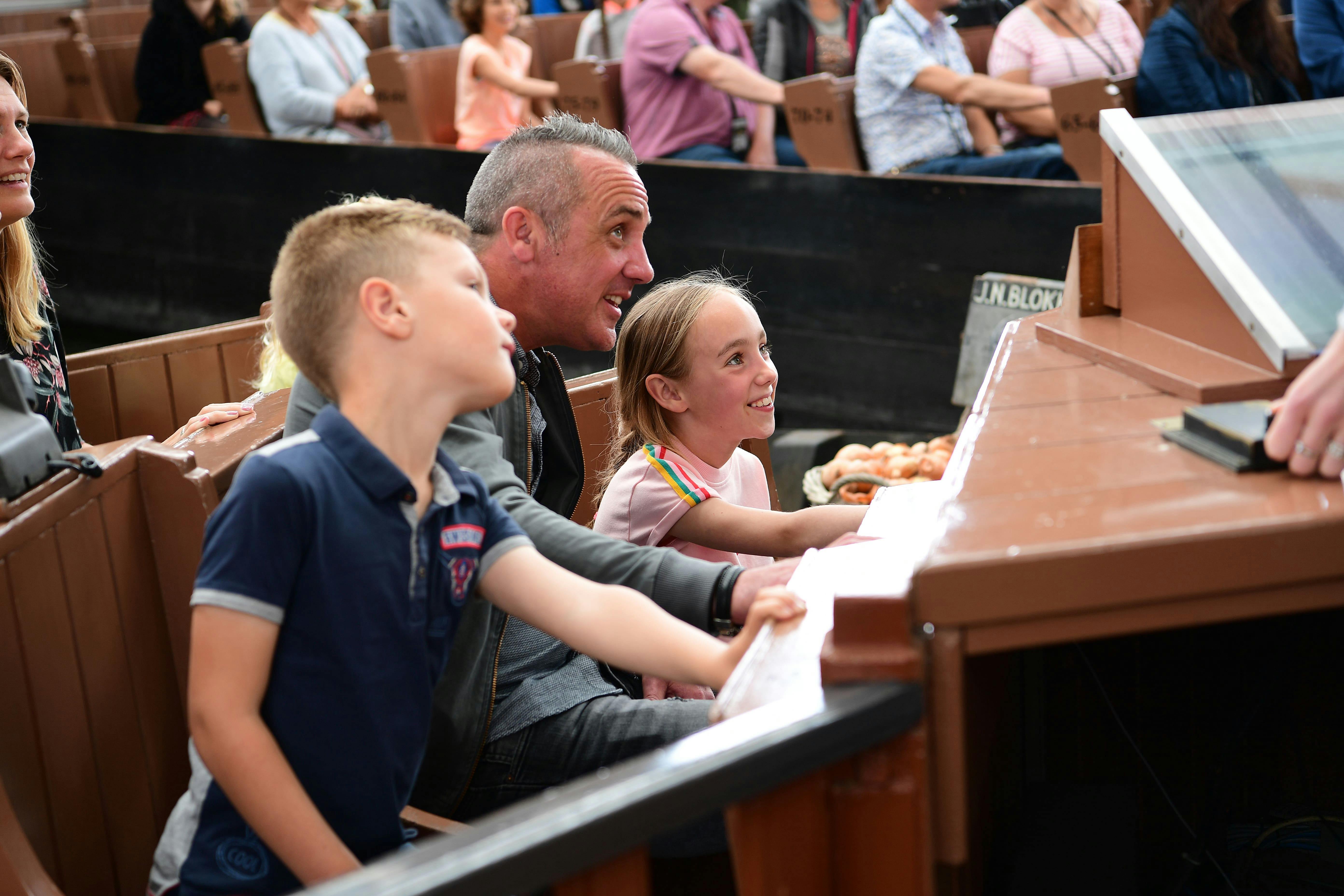 Ein Mann und zwei Kinder sitzen in einem Auto, lächelnd und beschäftigt, umgeben von anderen Menschen im Hintergrund.