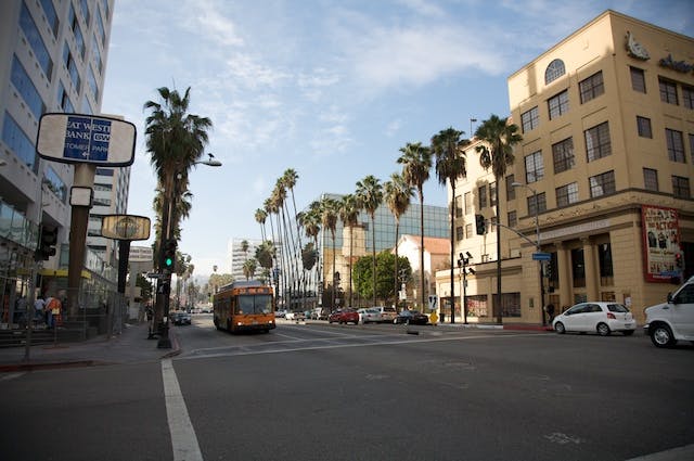 City street with palm trees, buildings, a bus, and cars, under a clear sky. Pedestrians walk along the sidewalks.
