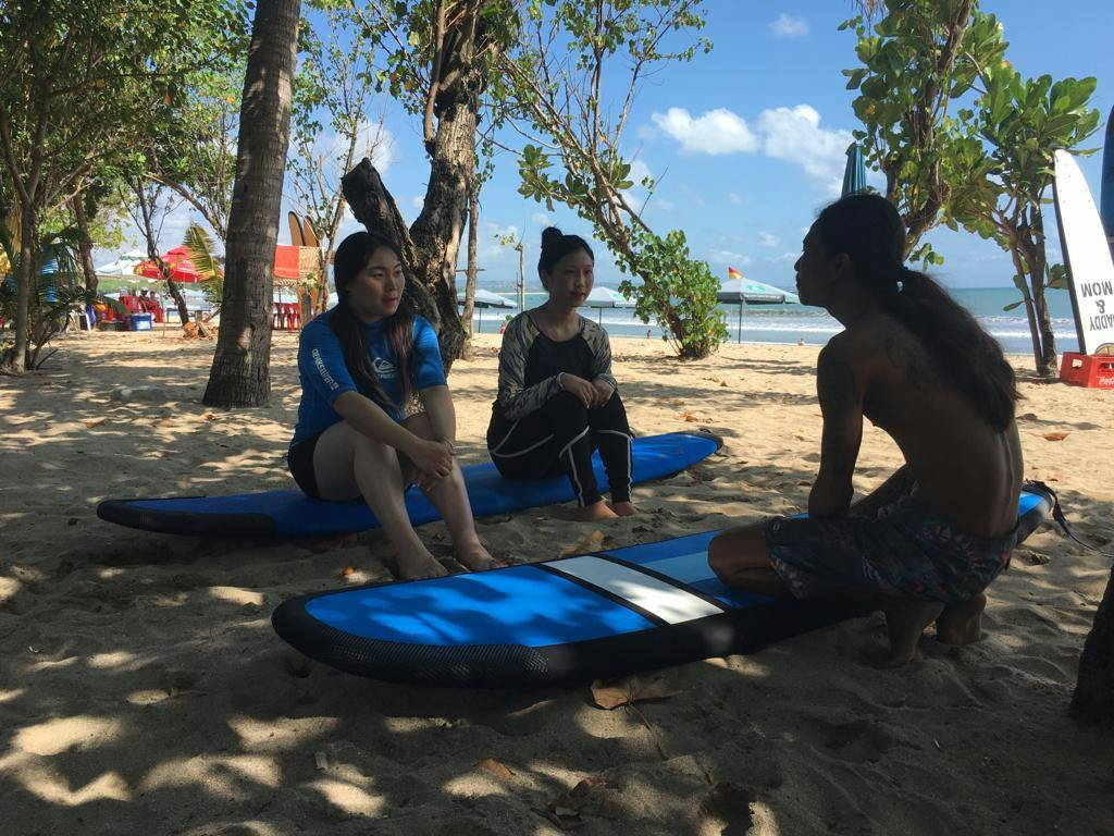 Trzy osoby siedzące na niebieskich deskach surfingowych na piaszczystej plaży pod drzewami, z widocznym morzem i łodziami w tle.