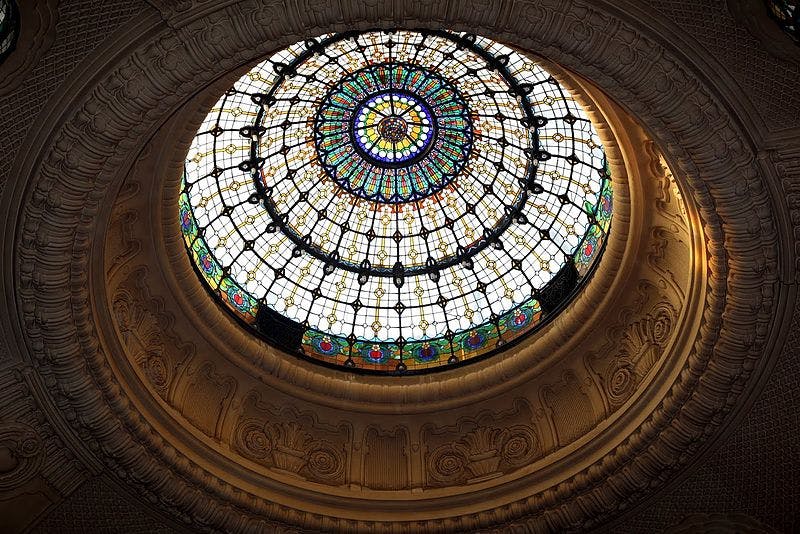 Kreisförmige Glaskuppel mit komplizierten Mustern und leuchtenden Farben, umgeben von kunstvollen architektonischen Details.