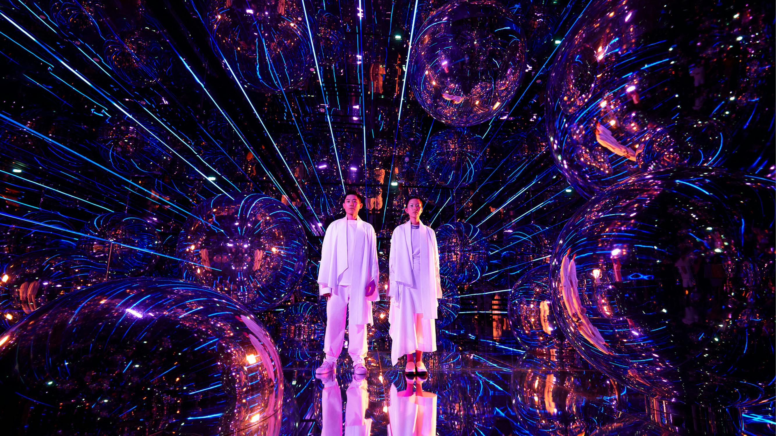 Δύο άτομα με λευκά ρούχα στέκονται σε ένα δωμάτιο γεμάτο με ανακλαστικές επιφάνειες, μεγάλες σφαίρες καθρέφτη και ζωντανά μοβ και μπλε φώτα.