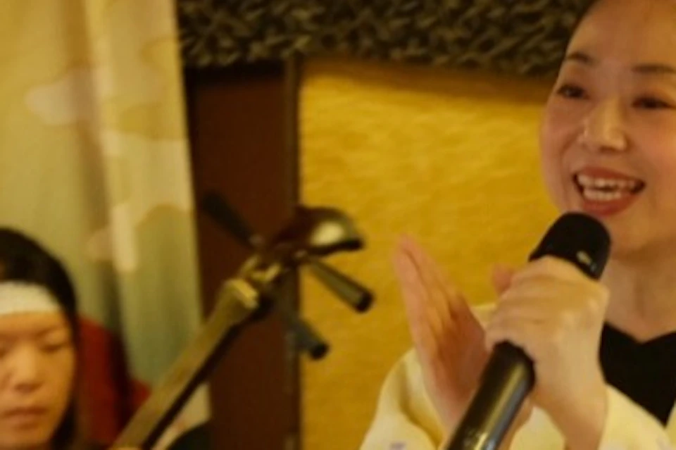 A woman in a yellow kimono sings into a microphone while a seated person plays a traditional string instrument. A mountain backdrop is behind them.