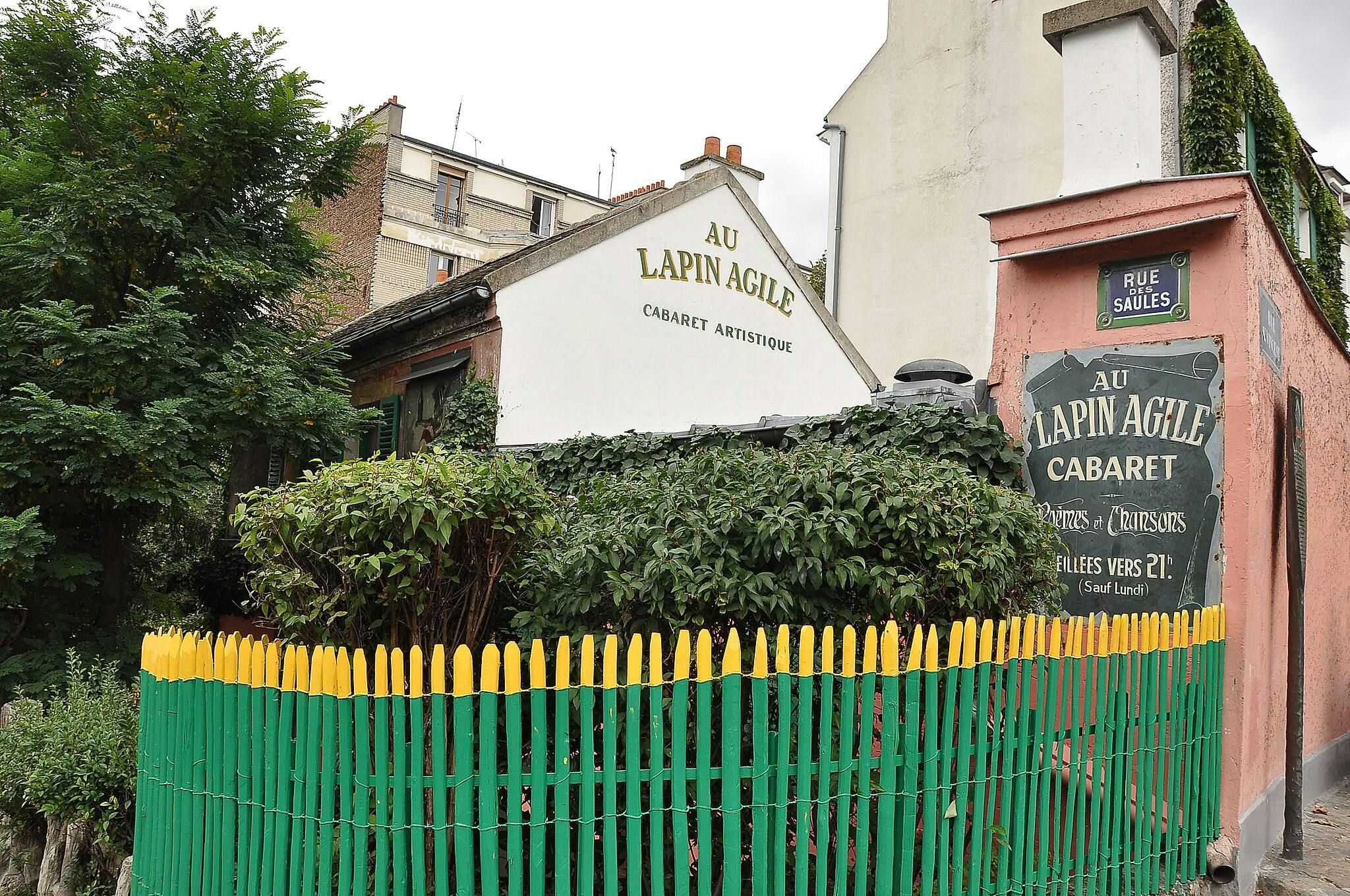 Ένα καλλιτεχνικό καμπαρέ που ονομάζεται "Au Lapin Agile" με πράσινο φύλλωμα, έναν πολύχρωμο φράχτη και μια πινακίδα σε έναν ροζ τοίχο κατά μήκος της Rue des Saules.