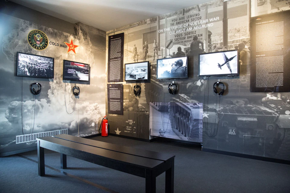 Museum exhibit with Cold War theme, featuring multimedia screens, headphones, informational panels, and a bench for visitors.