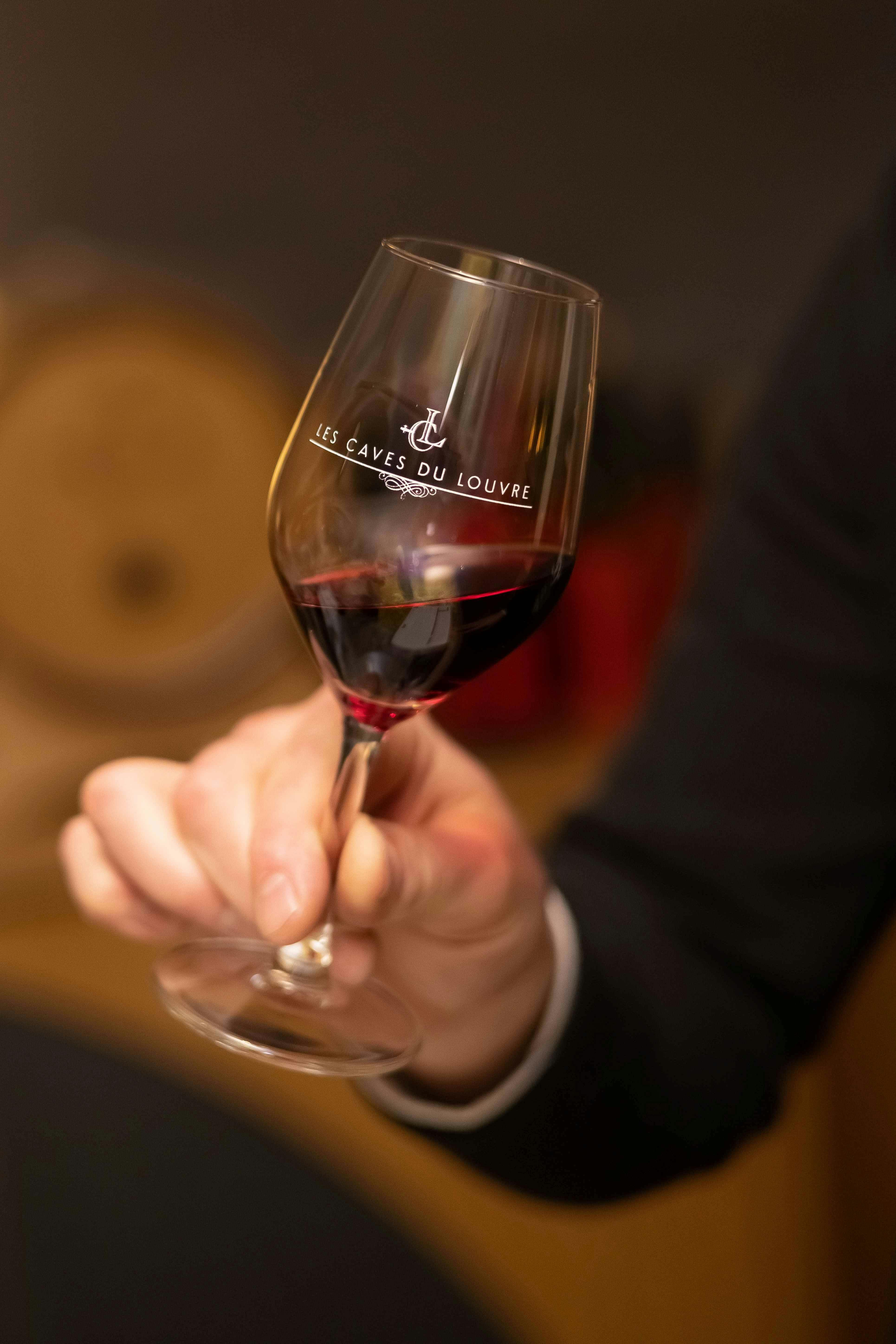 ぼやけた背景の「ルーブル美術館の洞窟」と書かれた赤ワインを詰めたワイングラスを手に持っています。