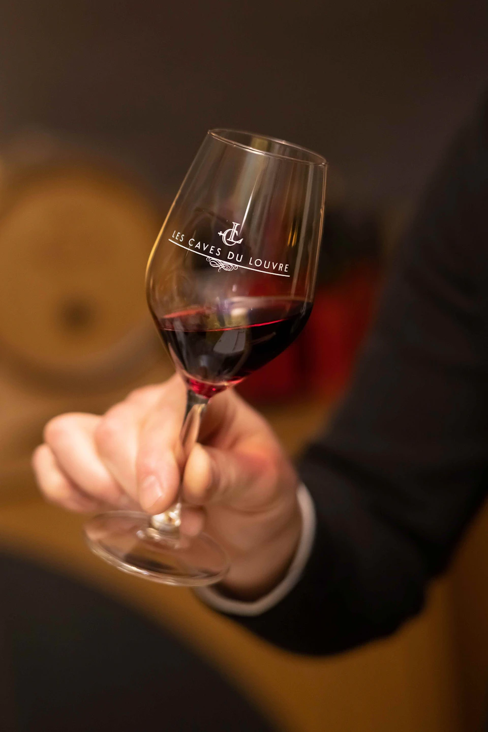 A hand holds a wine glass filled with red wine, labeled "Les Caves du Louvre," with a blurred background.