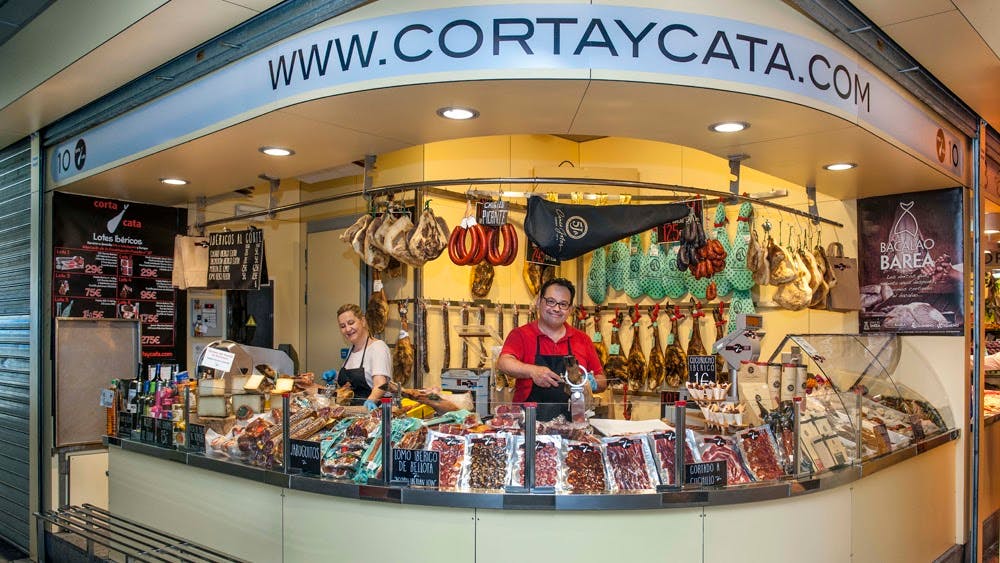 Lahůdkářský pult s masem a klobásami na displeji, obsazený dvěma usměvavými pracovníky, z nichž jeden krájí maso, v tržnici.