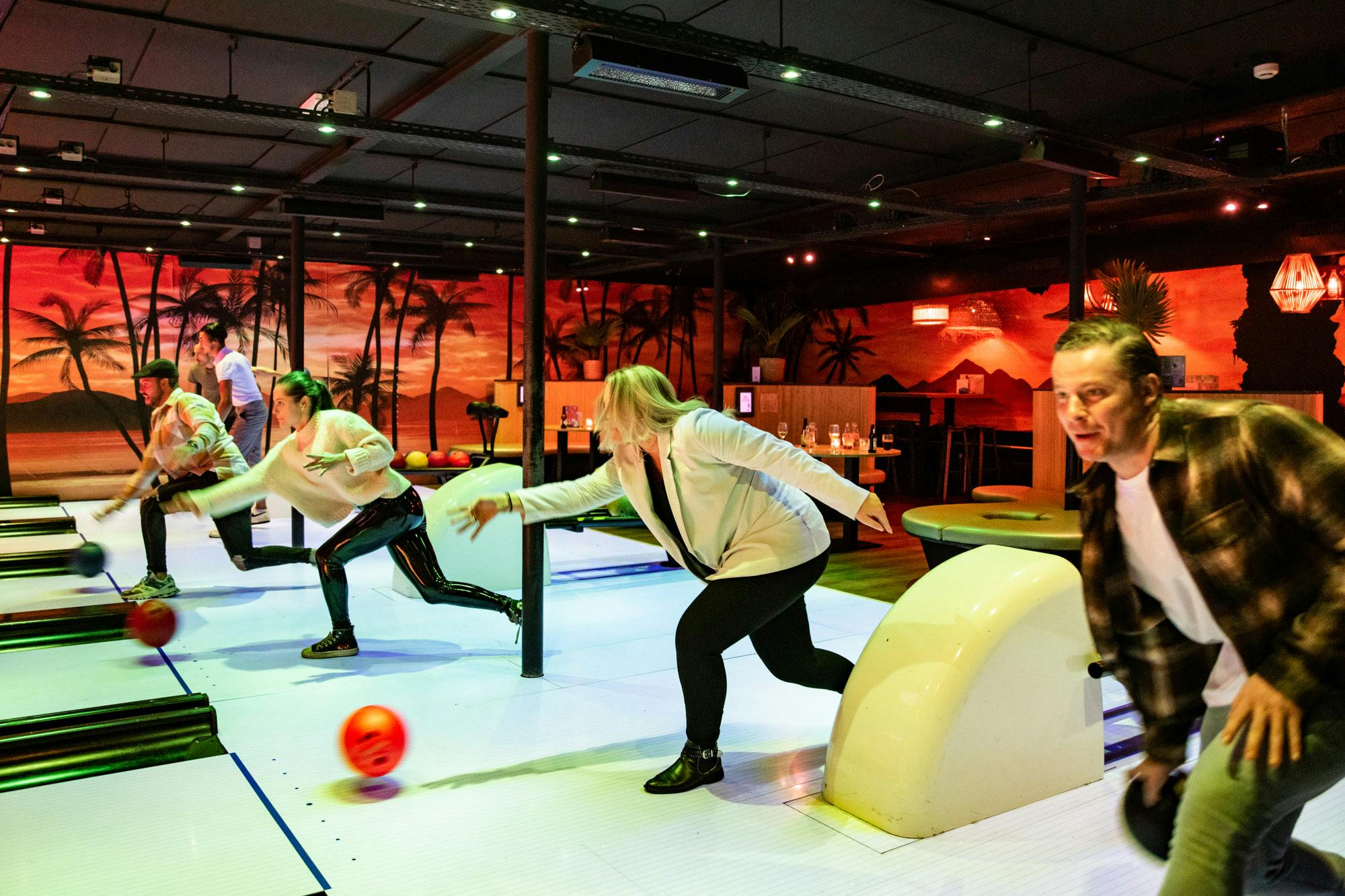 Τέσσερα άτομα σε μια αίθουσα μπόουλινγκ παίζουν ενεργά μπόουλινγκ, με μια τροπική τοιχογραφία στο παρασκήνιο.
