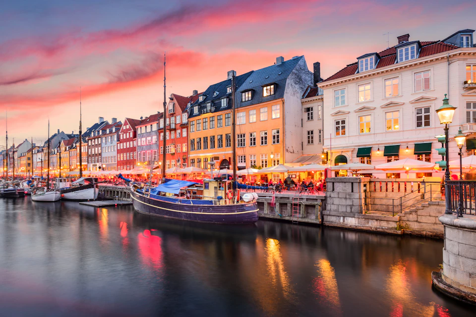 Waterside scene with colorful buildings, docked boats, and outdoor cafes under a pink and purple sunset sky.