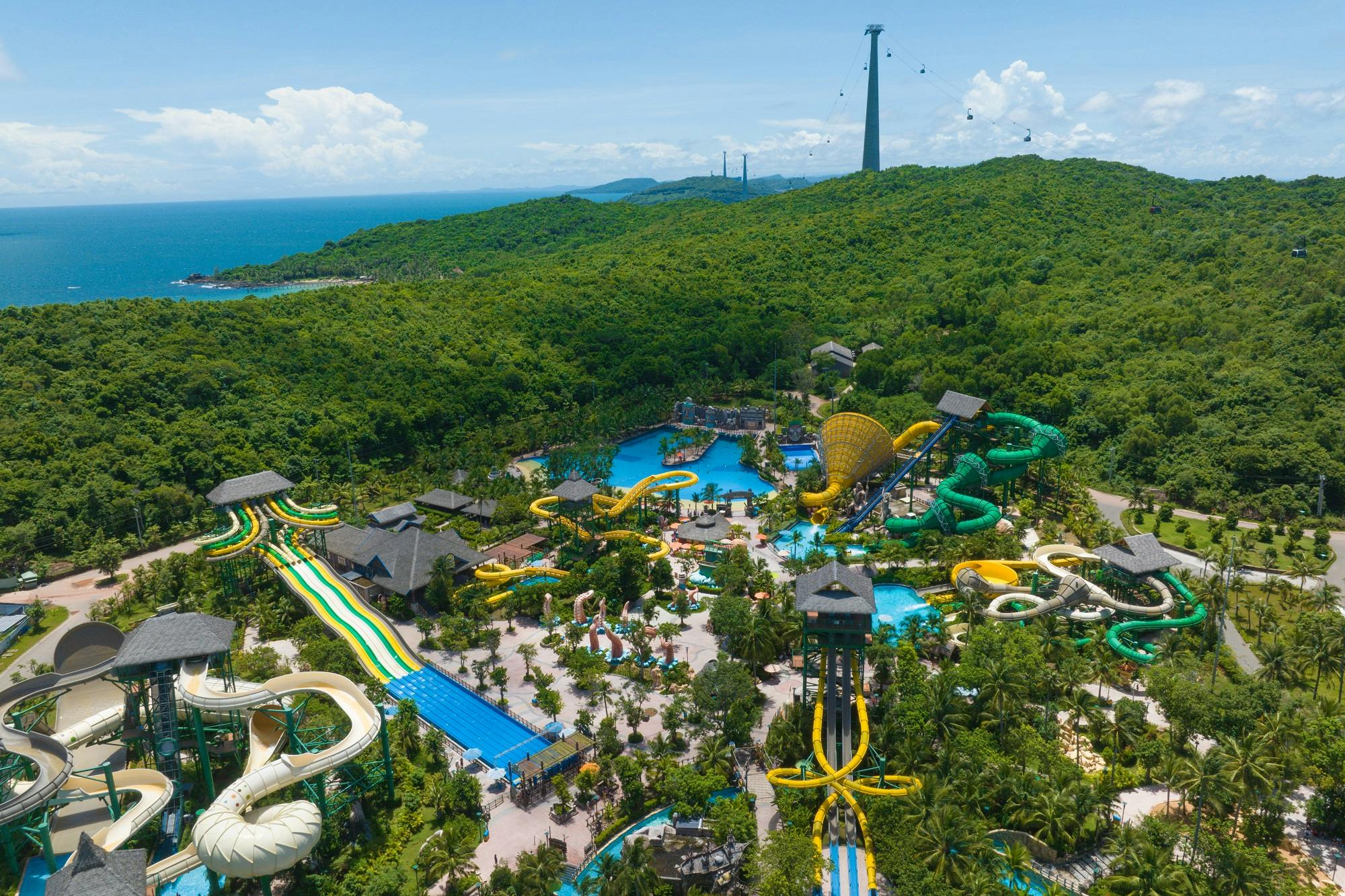 A large water park with colorful slides and pools surrounded by lush greenery and a cable car in the distance.