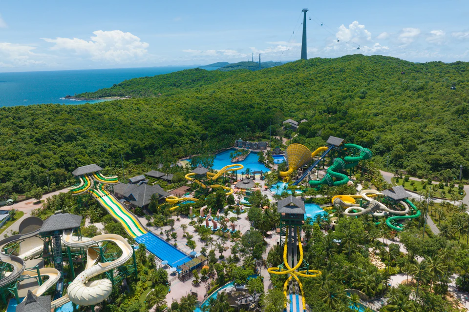 Aerial view of a water park with colorful slides, pools, and greenery, set in a lush forest near the sea.