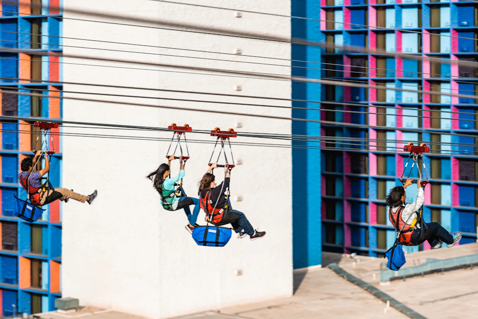 People zip-lining near a colorful building facade with blue, pink, and black accents.