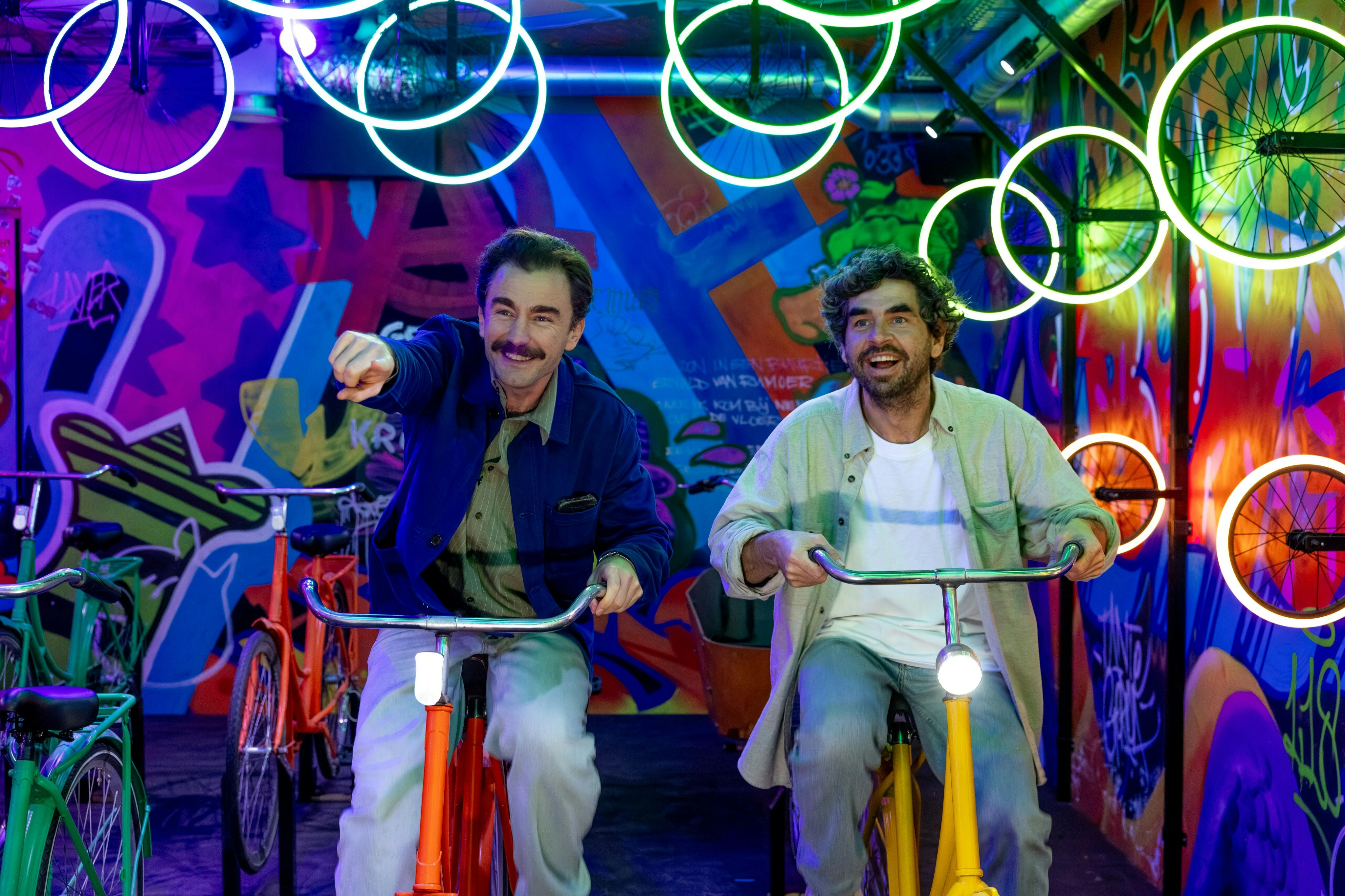 Δύο άνδρες με στατικά ποδήλατα σε ένα πολύχρωμο δωμάτιο με φώτα νέον και γκράφιτι.