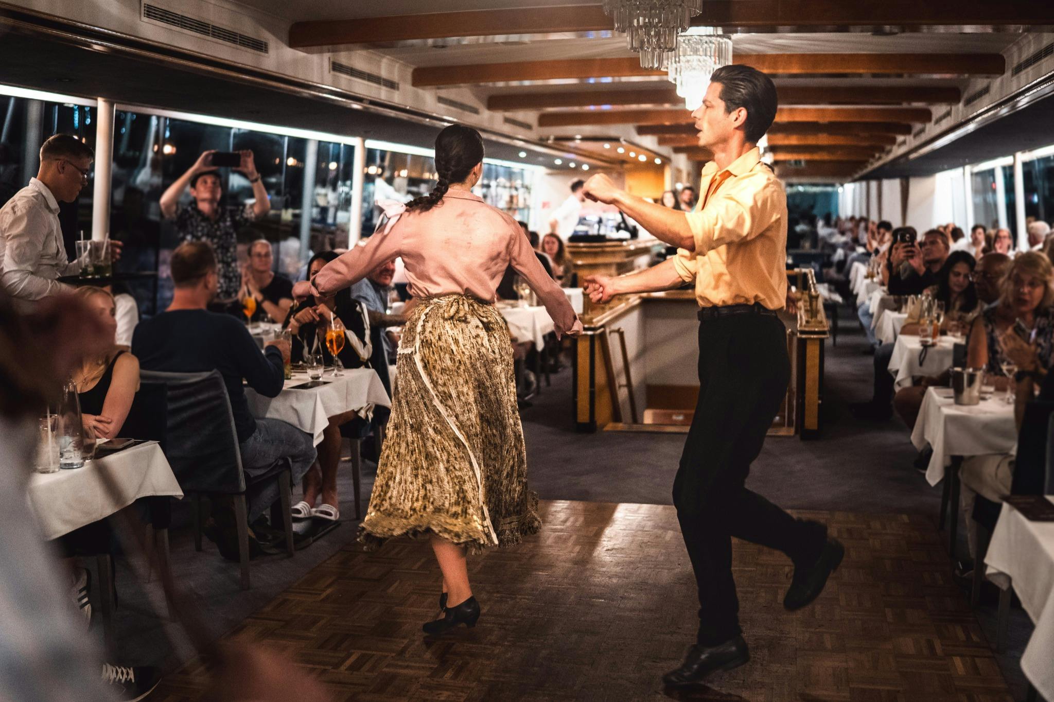 Ein Paar tanzt in einem Restaurant mit sitzenden Gästen, polierten Böden, elegantem Dekor und einer lebhaften Atmosphäre.
